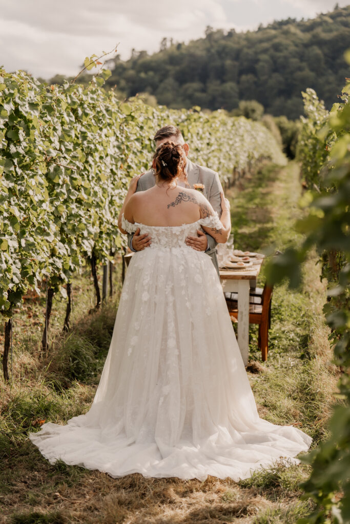 Braut und Bräutigam umarmen sich in einem Weinberg, hinter ihnen steht ein Tisch auf einem Grasweg.