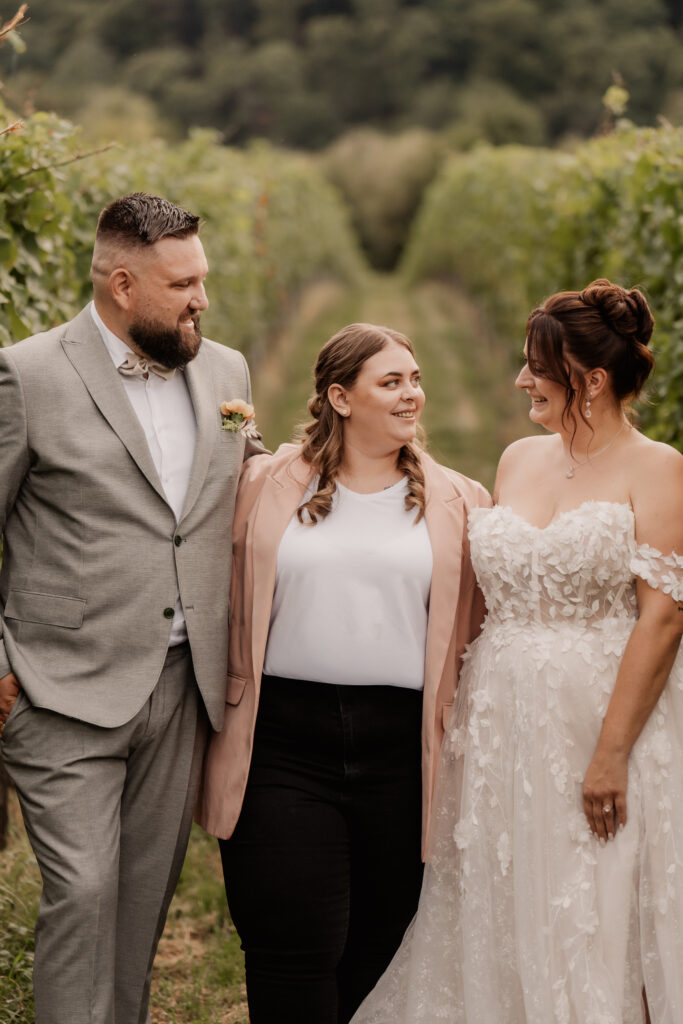 Ein lächelndes Paar mit seiner Hochzeitsplanerin, die Frau steht zwischen ihnen in einem Weinberg, während die Braut ein schönes weißes Kleid trägt.