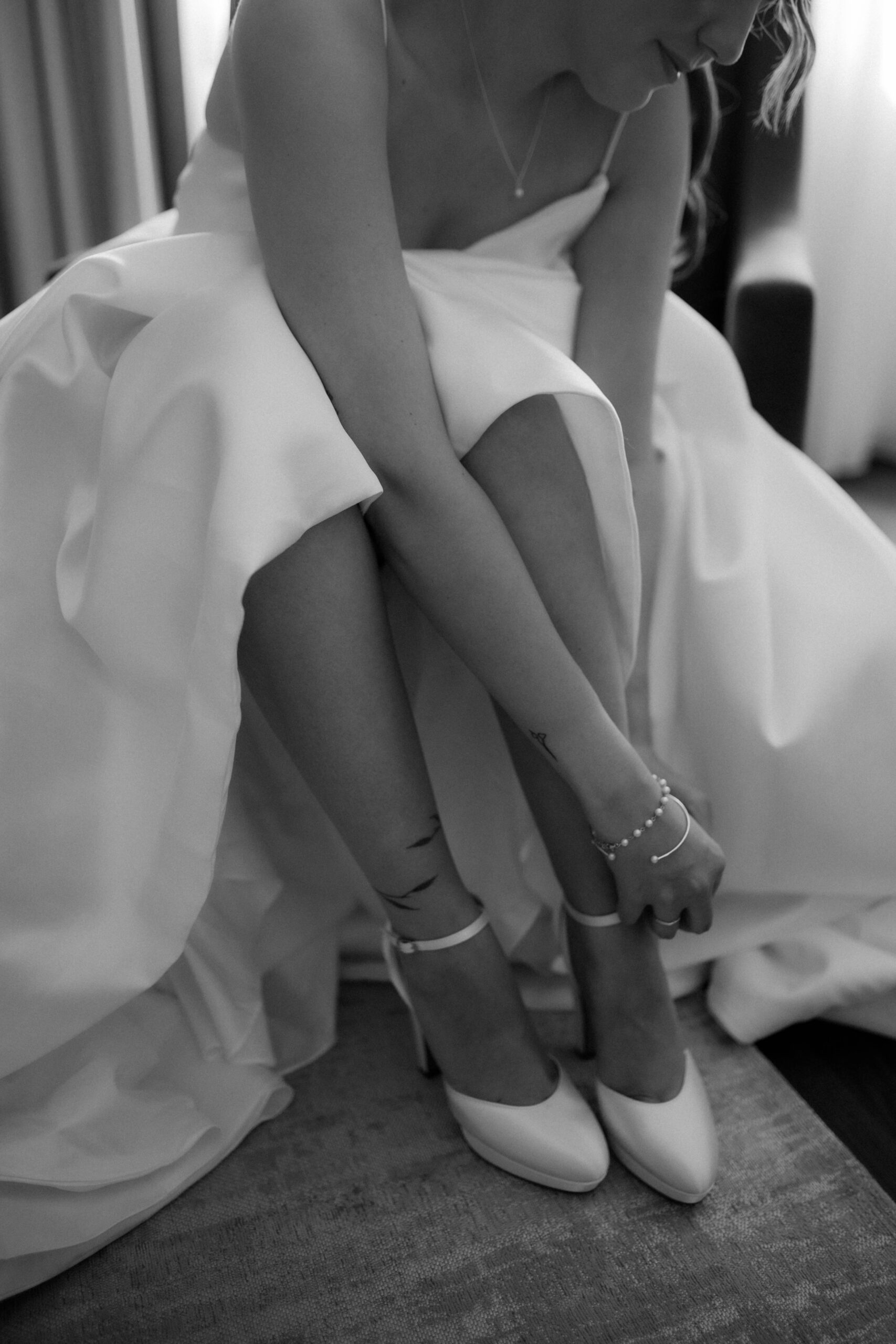 Braut im Hochzeitskleid sitzend und ein Knöchelriemchen an ihrem hochhackigen Schuh befestigend, perfekt einen ruhigen Moment der Hochzeitsplanung in Rheinland-Pfalz einfangend.