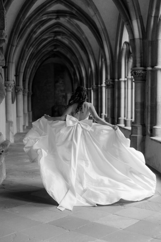 Eine Braut in einem wallenden Kleid mit Schleife schreitet, geführt von ihrer Hochzeitsplanerin, anmutig durch einen steinernen Bogengang.