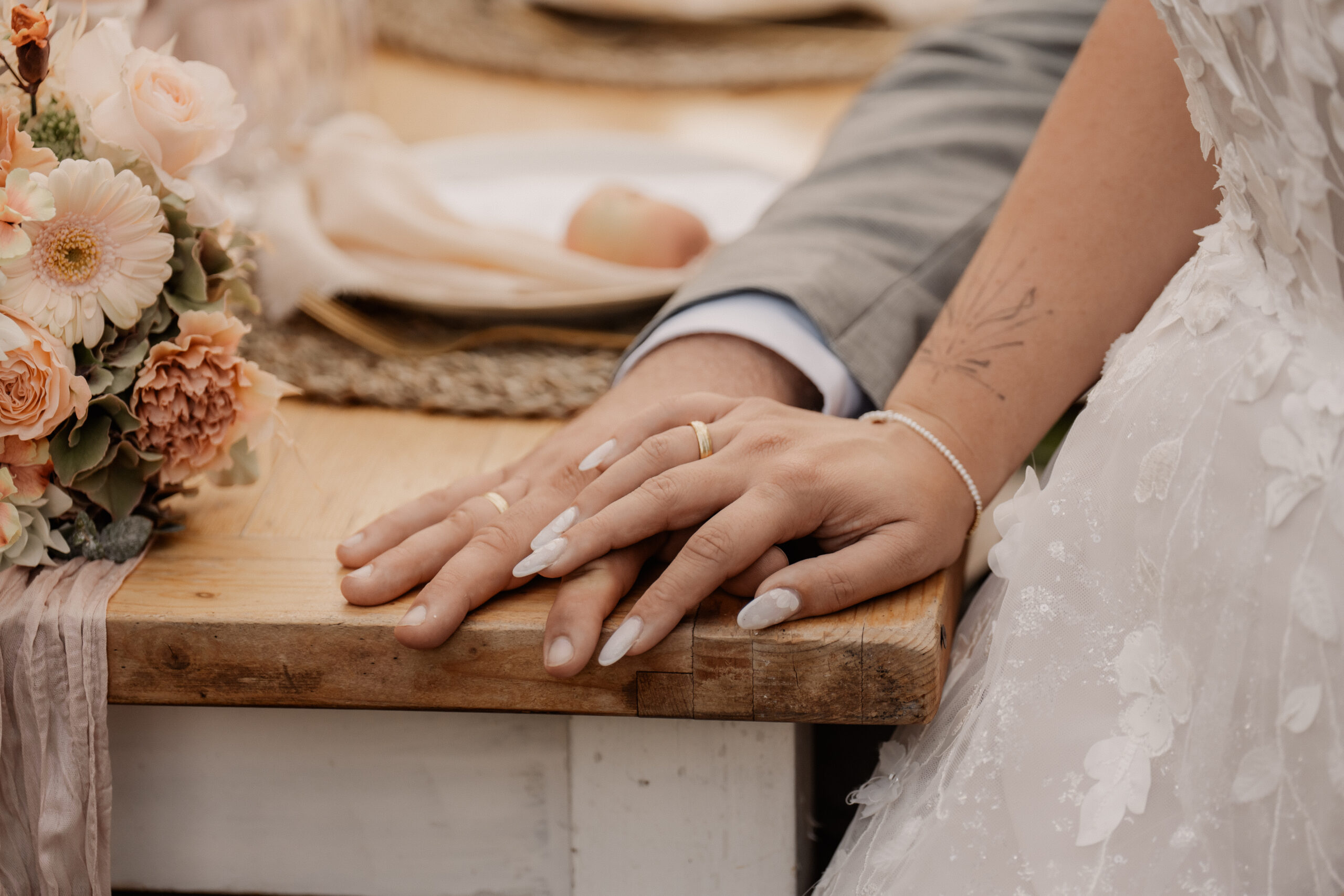 Eine Braut und ein Bräutigam legen ihre Hände mit den Eheringen auf einen Holztisch neben einem Blumenstrauß, der von einer Hochzeitsplanerin aus Trier wunderschön arrangiert wurde.