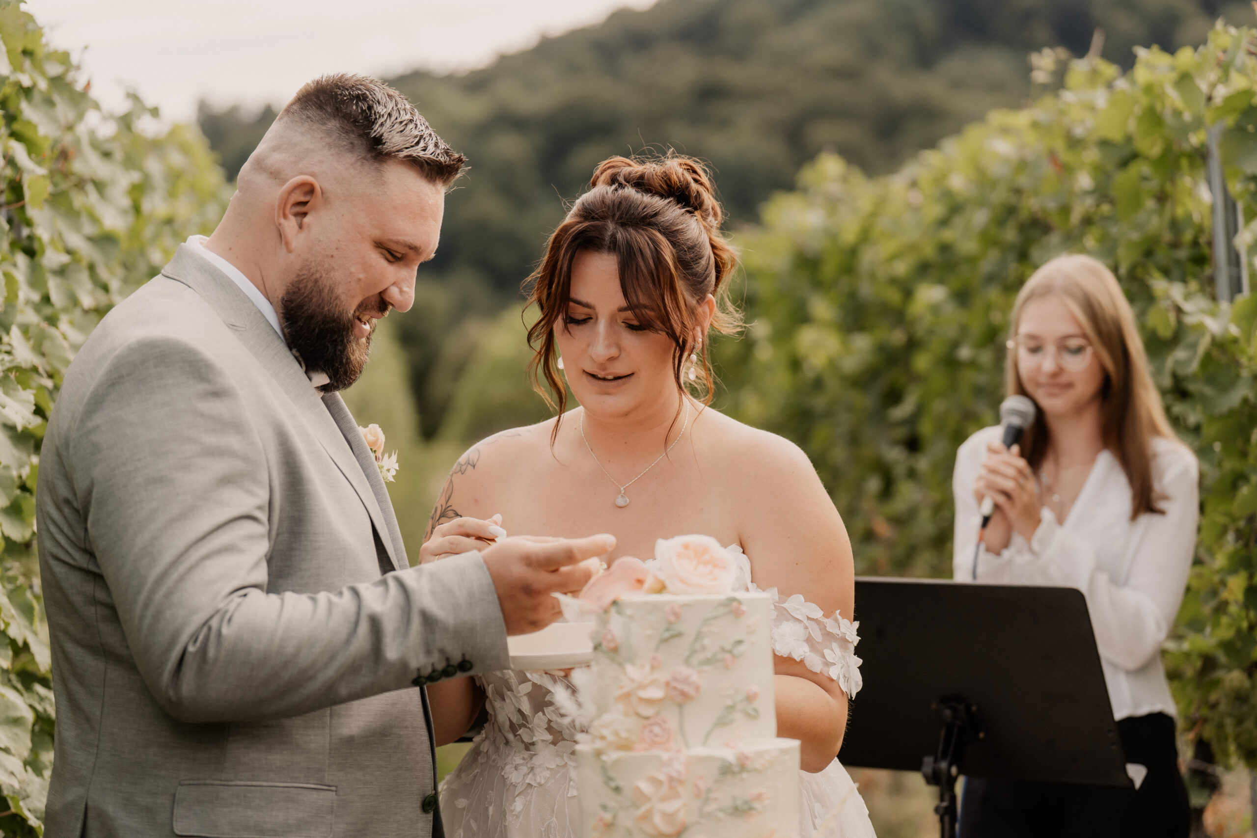 Braut und Bräutigam schneiden ihre Hochzeitstorte im Freien in Trier an, während eine Hochzeitsplanerin hinter ihnen in ein Mikrofon spricht.