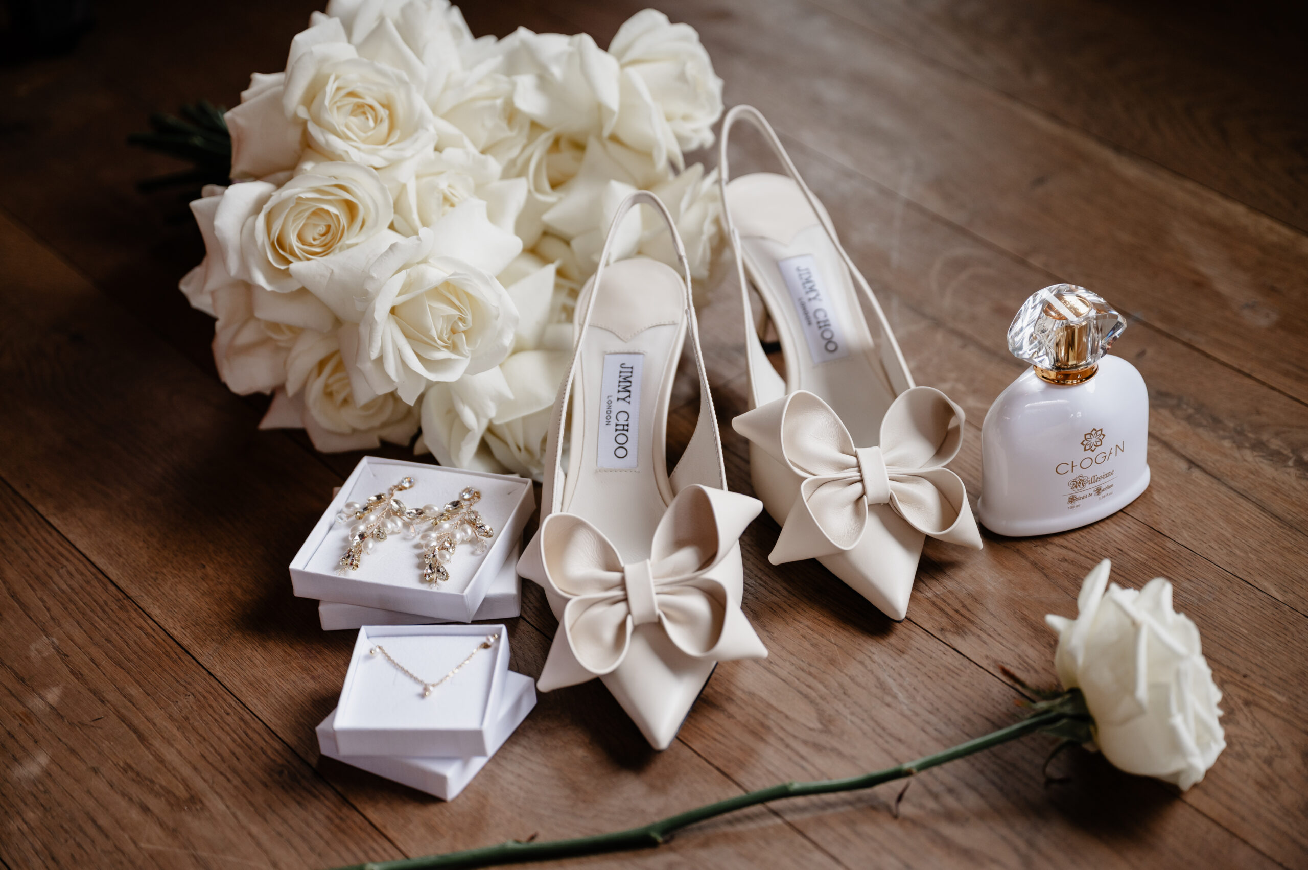 Cremefarbene Absätze mit Schleifen, weiße Rosen, Perlenschmuck und eine Parfümflasche auf einem Holzboden - die perfekte Inspiration für eine Hochzeitsplanerin im Saarland oder in Trier.