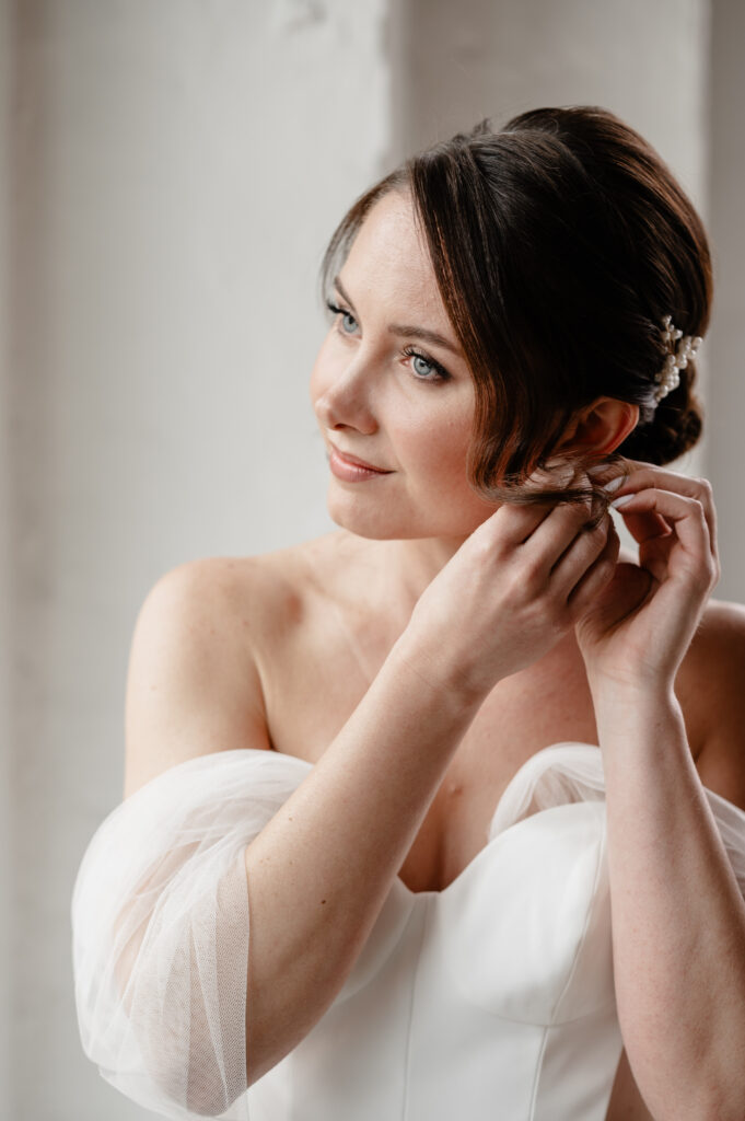 Eine Braut in einem weißen Kleid lächelt sanft, während sie sich einen Ohrring ansteckt, und hält damit einen magischen Moment der Hochzeitsplanung in Trier, Rheinland-Pfalz, fest.