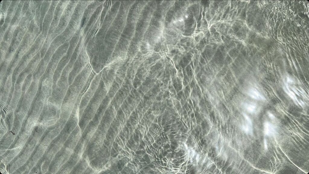 Das Sonnenlicht erzeugt auf dem flachen, klaren Wasser über sandigen Wellen am Grund wellenförmige Muster.