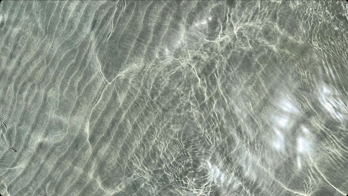 Das Sonnenlicht erzeugt auf dem flachen, klaren Wasser über sandigen Wellen am Grund wellenförmige Muster.