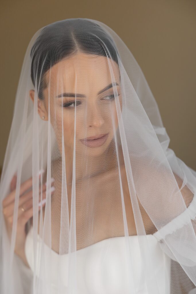 Eine Braut in weißem Kleid und Schleier blickt zur Seite vor einem weichen, neutralen Hintergrund, perfekt für eine romantische Hochzeit im Saarland oder in Rheinland-Pfalz.
