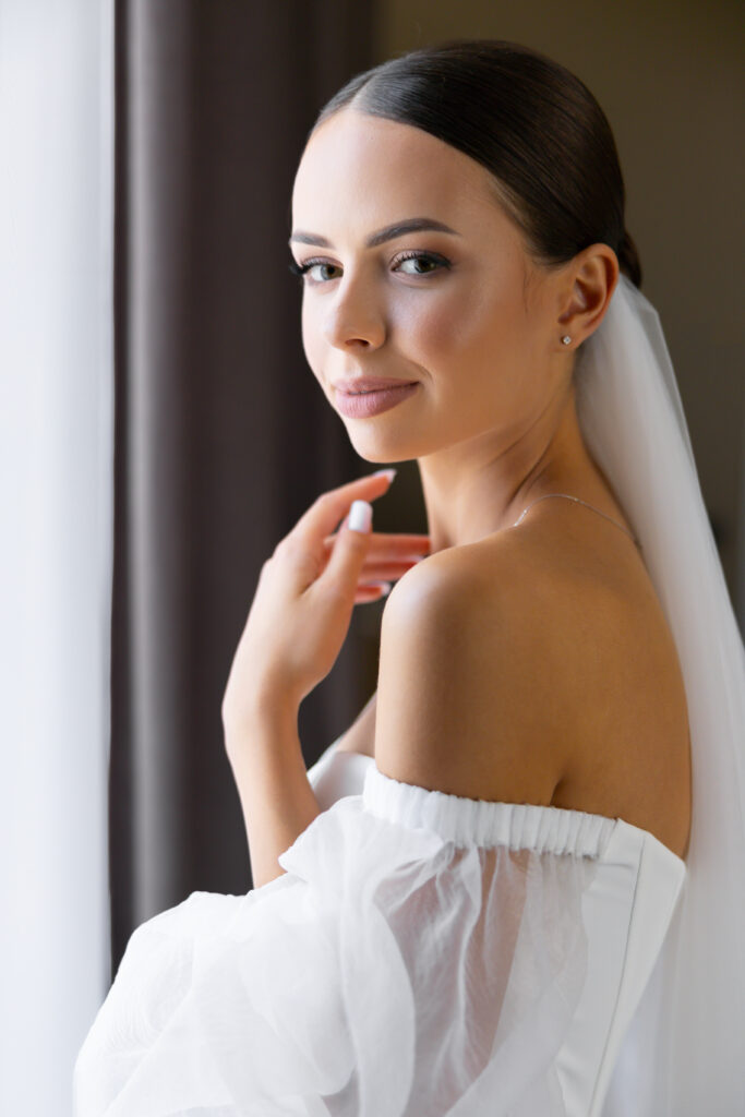 Eine Braut in einem schulterfreien weißen Kleid und Schleier steht an einem Fenster im Saarland, schaut über ihre Schulter und lächelt, um einen perfekten Moment der Hochzeitsplanung festzuhalten.