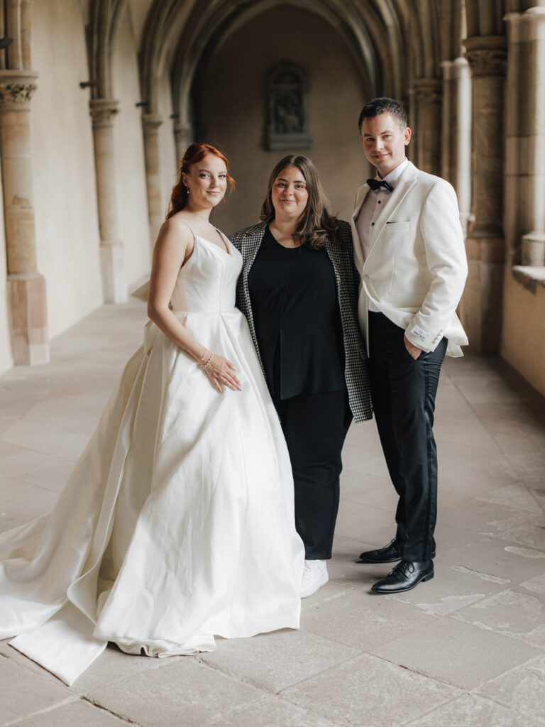 Eine Braut, ein Bräutigam und ihre Hochzeitsplanerin stehen zusammen in einem Flur mit gewölbter Decke und lächeln in die Kamera.