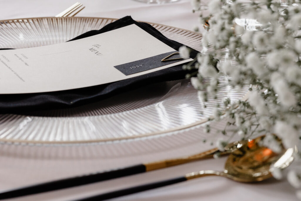 Eine Nahaufnahme eines elegant gedeckten Tisches mit einer Speisekarte auf einer Glasplatte, goldenem Besteck und weißen Blumen.