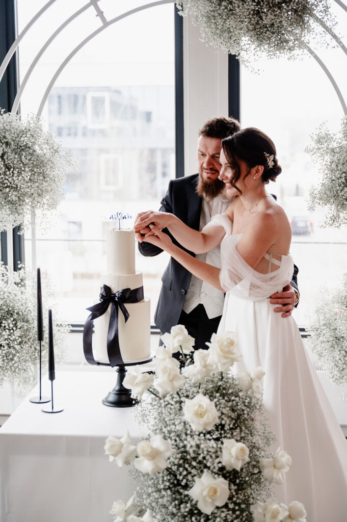 Eine Braut und ein Bräutigam schneiden gemeinsam eine weiße Hochzeitstorte an, umgeben von weißen Blumen und schwarzen Kerzen.