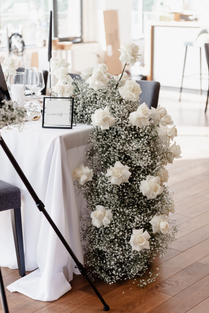 Weiße Rosen und Schleierkraut am Rande eines elegant gedeckten Tisches bei einer Indoor-Veranstaltung.