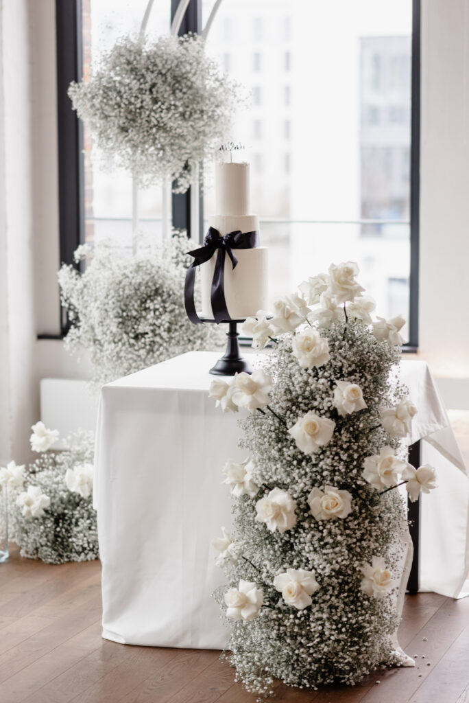 Eine weiße Hochzeitstorte auf einem Ständer, umgeben von weißen Rosen und Blumenarrangements mit Babyatem.