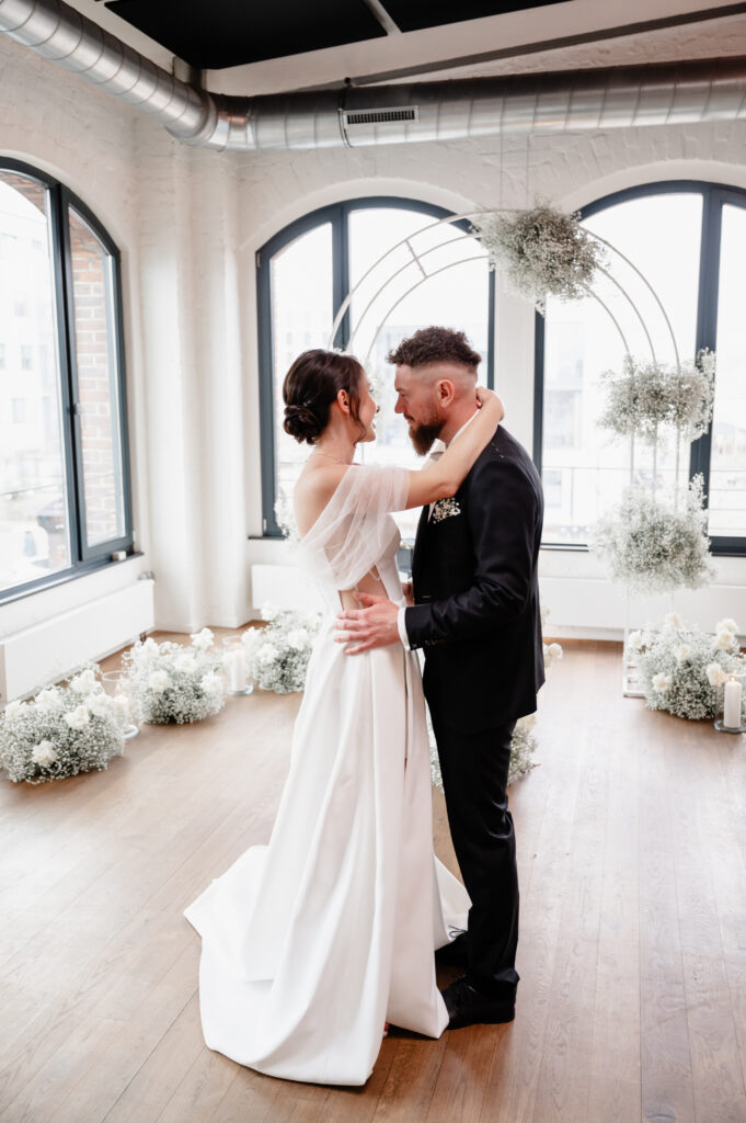 Eine Braut und ein Bräutigam tanzen zusammen in einem hellen Raum, der mit weißen Blumen und großen Fenstern geschmückt ist.