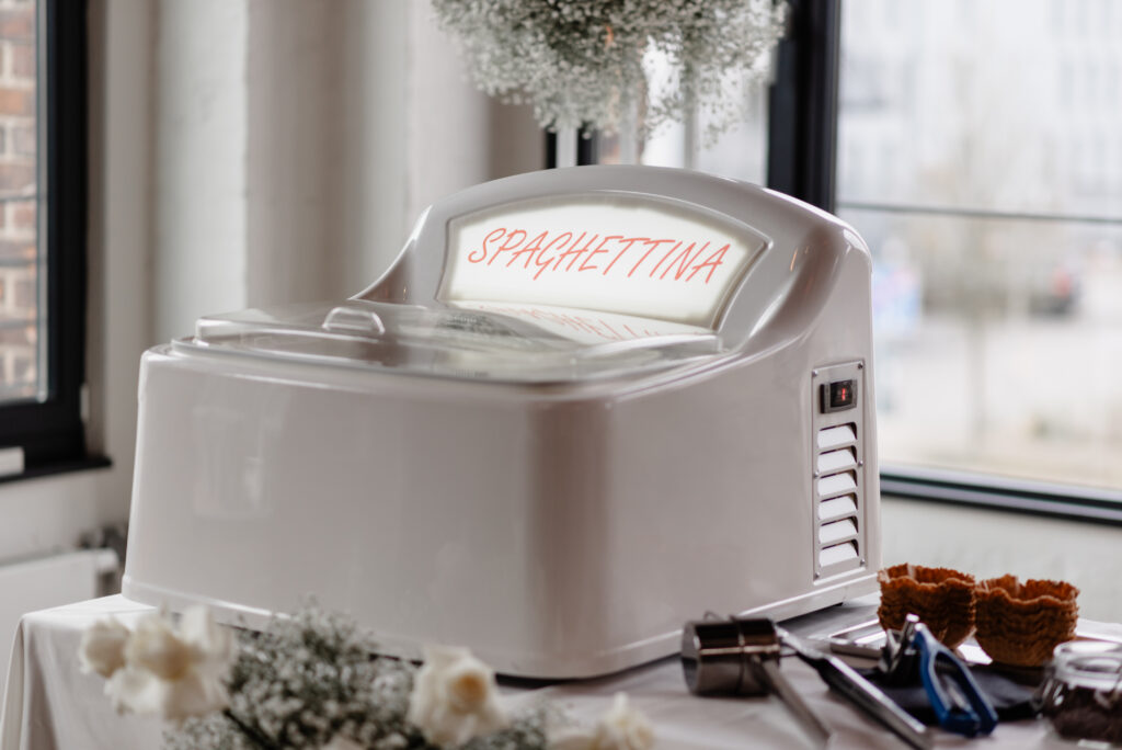 Eine weiße Maschine mit der Aufschrift SPAGHETTINA auf einem Tisch, daneben Blumen, Tüten und Servierutensilien.