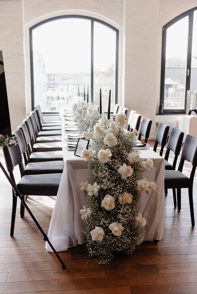 Elegant gedeckter Tisch für eine Veranstaltung mit weißen Rosen und einem Blumenarrangement mit Babyatem in der Nähe großer Fenster.