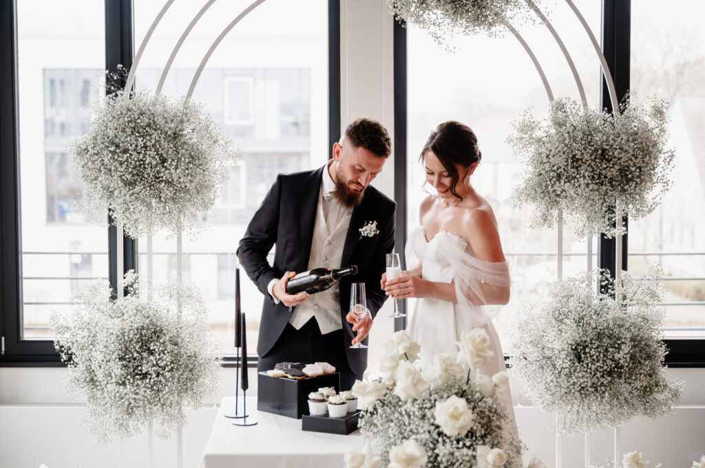 Braut und Bräutigam stehen an einem Tisch mit Blumen, während der Bräutigam Champagner in ein Glas schüttet.
