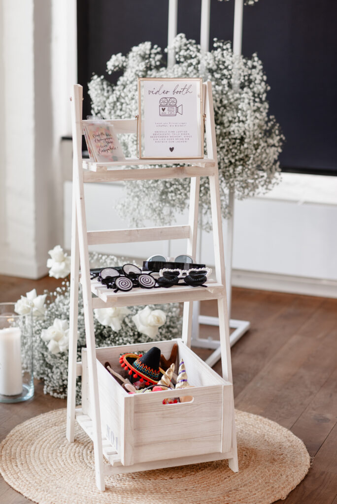 Weißes Holzregal mit Fotokabinenrequisiten, Blumen und einem gerahmten Schild obenauf, auf einem gewebten Teppich aufgestellt.