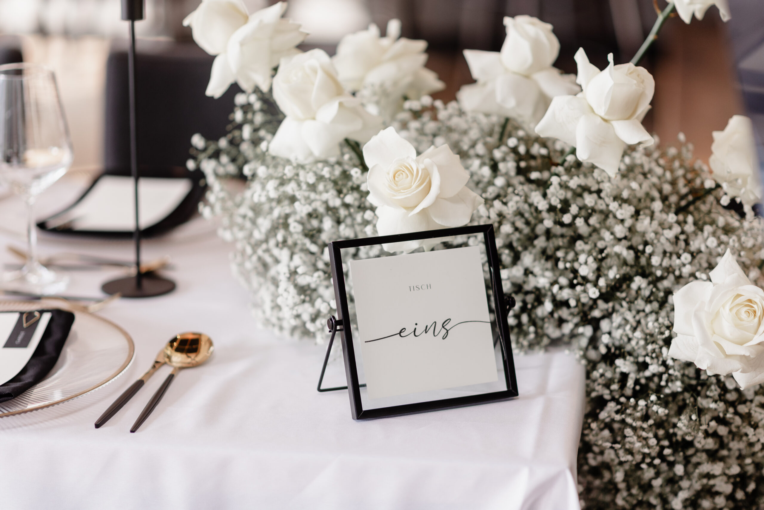 Elegant gedeckter Tisch mit weißen Rosen, Schleierkraut und einem gerahmten Schild mit der Aufschrift TISCH eins auf einem weißen Tuch.