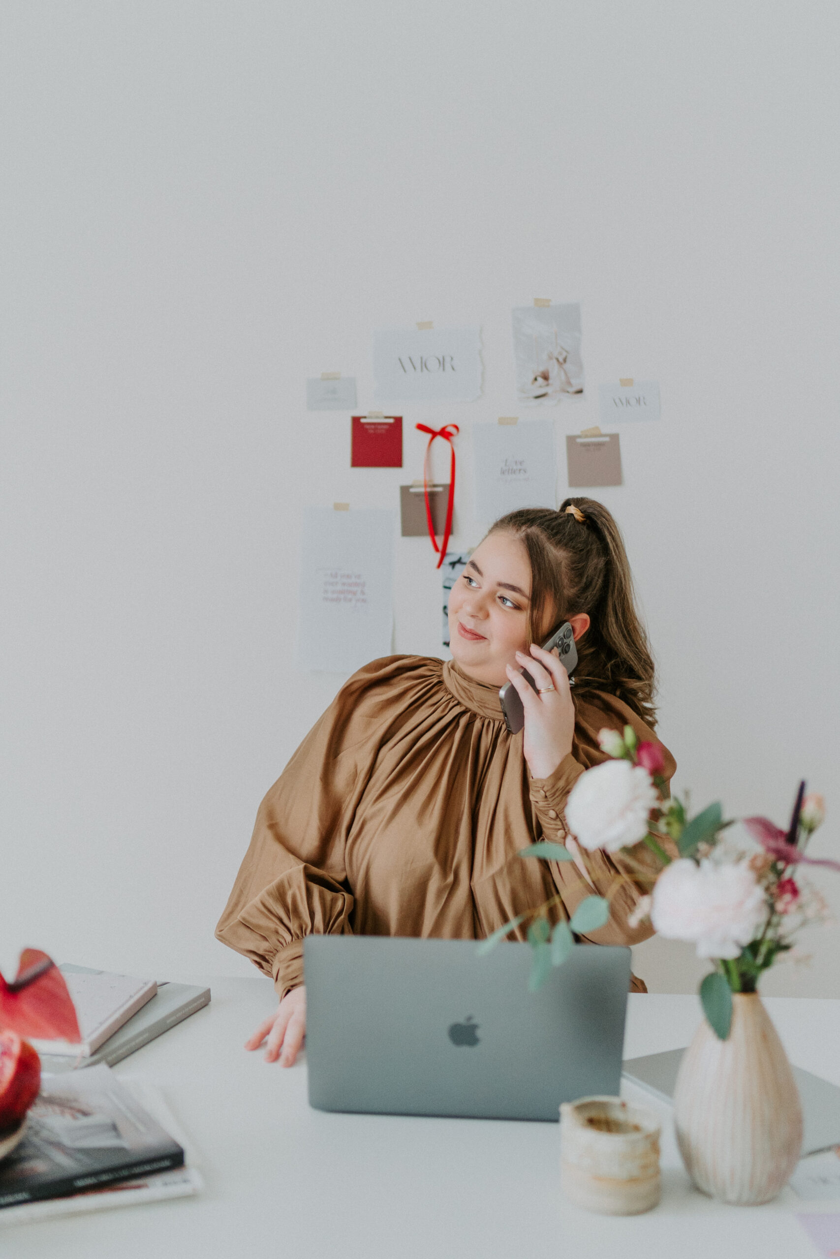 Frau in brauner Bluse lächelt, während sie an einem Schreibtisch mit Laptop und Blumen telefoniert.
