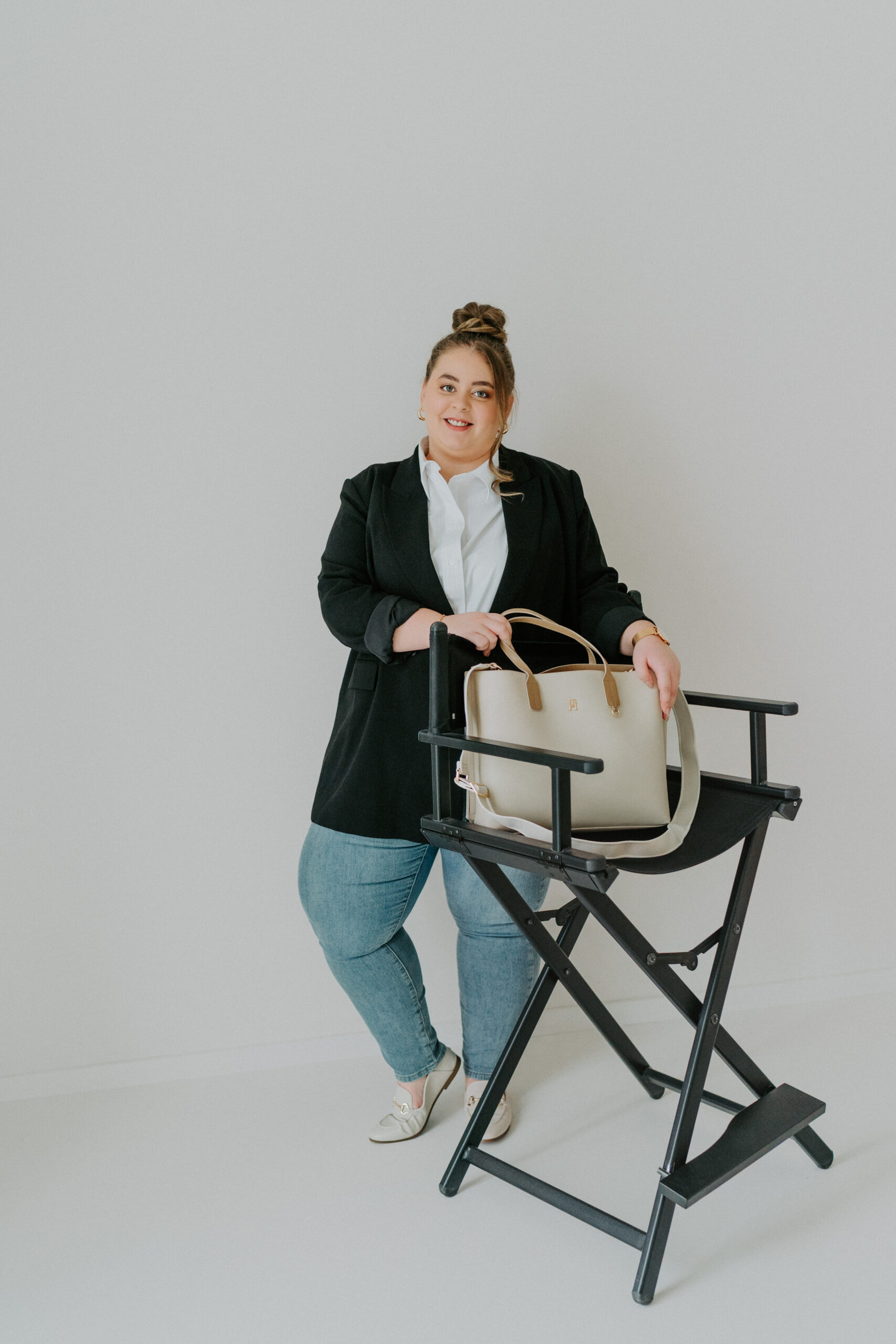 Eine Frau in Blazer und Jeans steht neben einem Regiestuhl, hält eine beigefarbene Handtasche und lächelt.