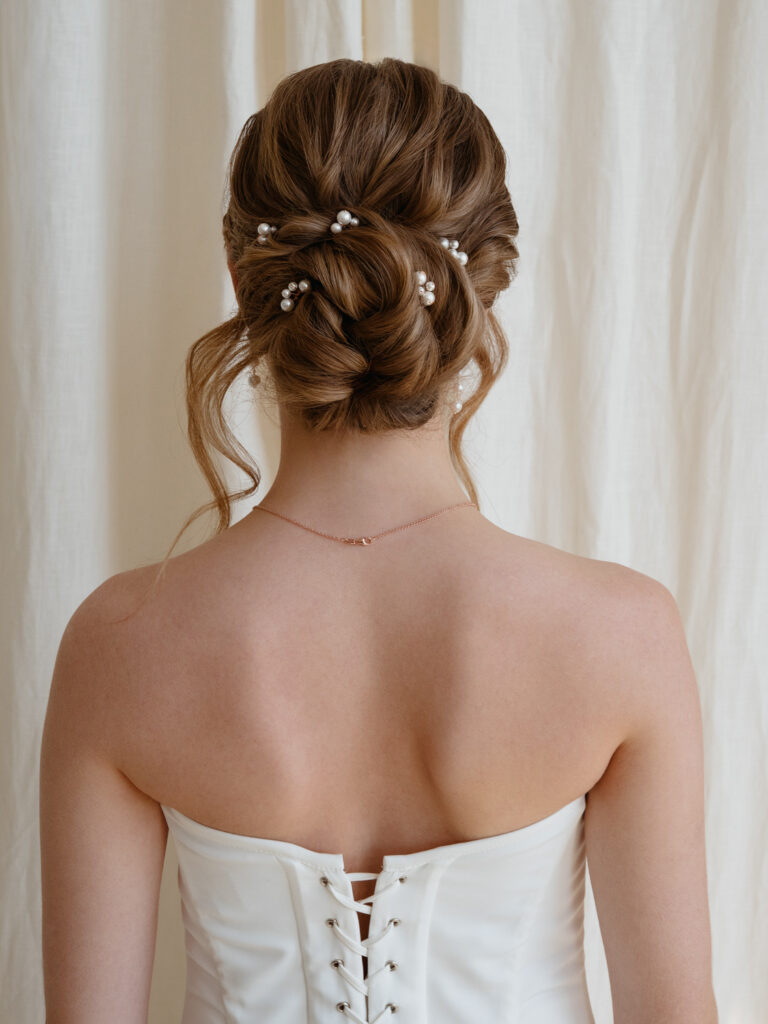 Eine Frau in einem weißen Schnürkleid mit einer eleganten Hochsteckfrisur, die mit kleinen Perlen verziert ist, von hinten gesehen.