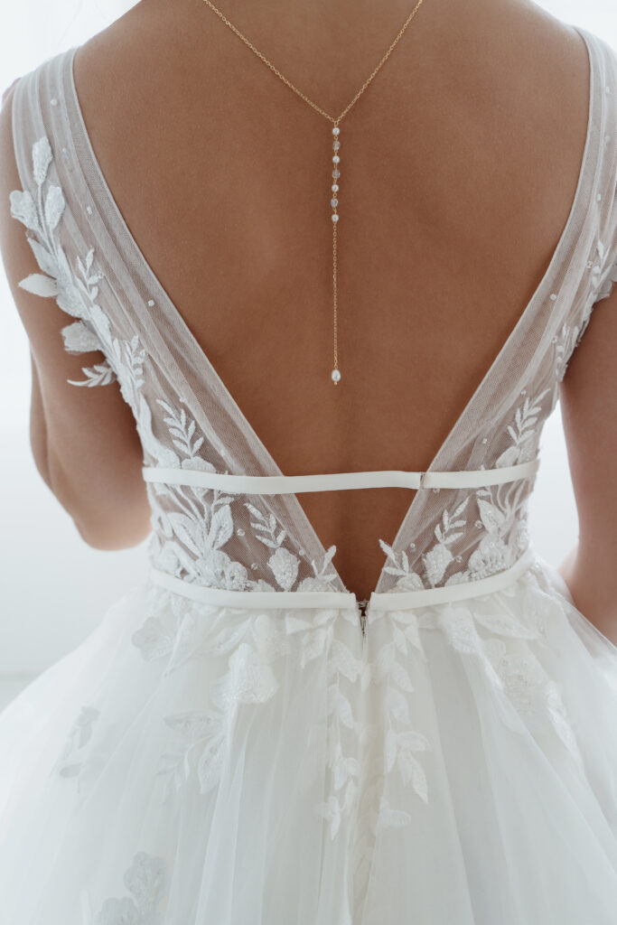 Braut in einem Spitzenhochzeitskleid mit tiefem V-Ausschnitt am Rücken, transparenten Blumenverzierungen und einer zarten Halskette am Rücken.