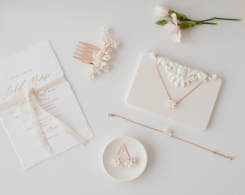 Flatlay-Aufnahme einer Hochzeitseinladung, eines Blumenschmuck-Sets, eines Kamms und rosa Blumen auf einer weißen Unterlage.