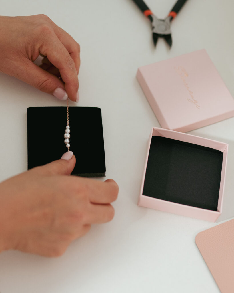 Hände legen eine Perlenkette in eine schwarze Schmuckschatulle, die neben einer rosa Verpackung und einer Drahtschere auf einem weißen Tisch steht.